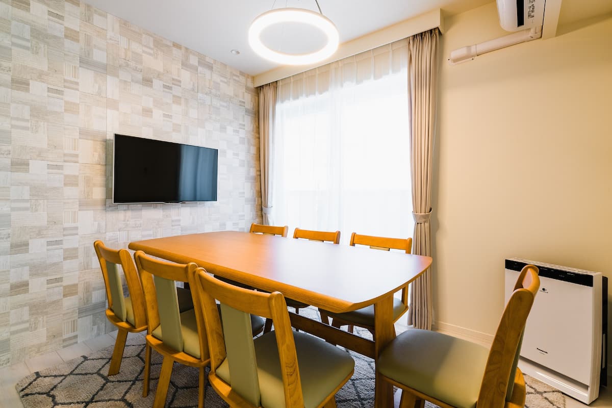 A dining area with a wooden table and chairs, a flat-screen TV mounted on the wall, and a window with curtains.