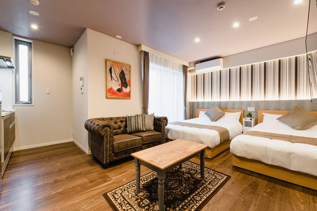 A modern hotel room with a wooden floor, a leather sofa, a wooden coffee table, two beds with white bedding, a nightstand, and a wall-mounted light fixture.