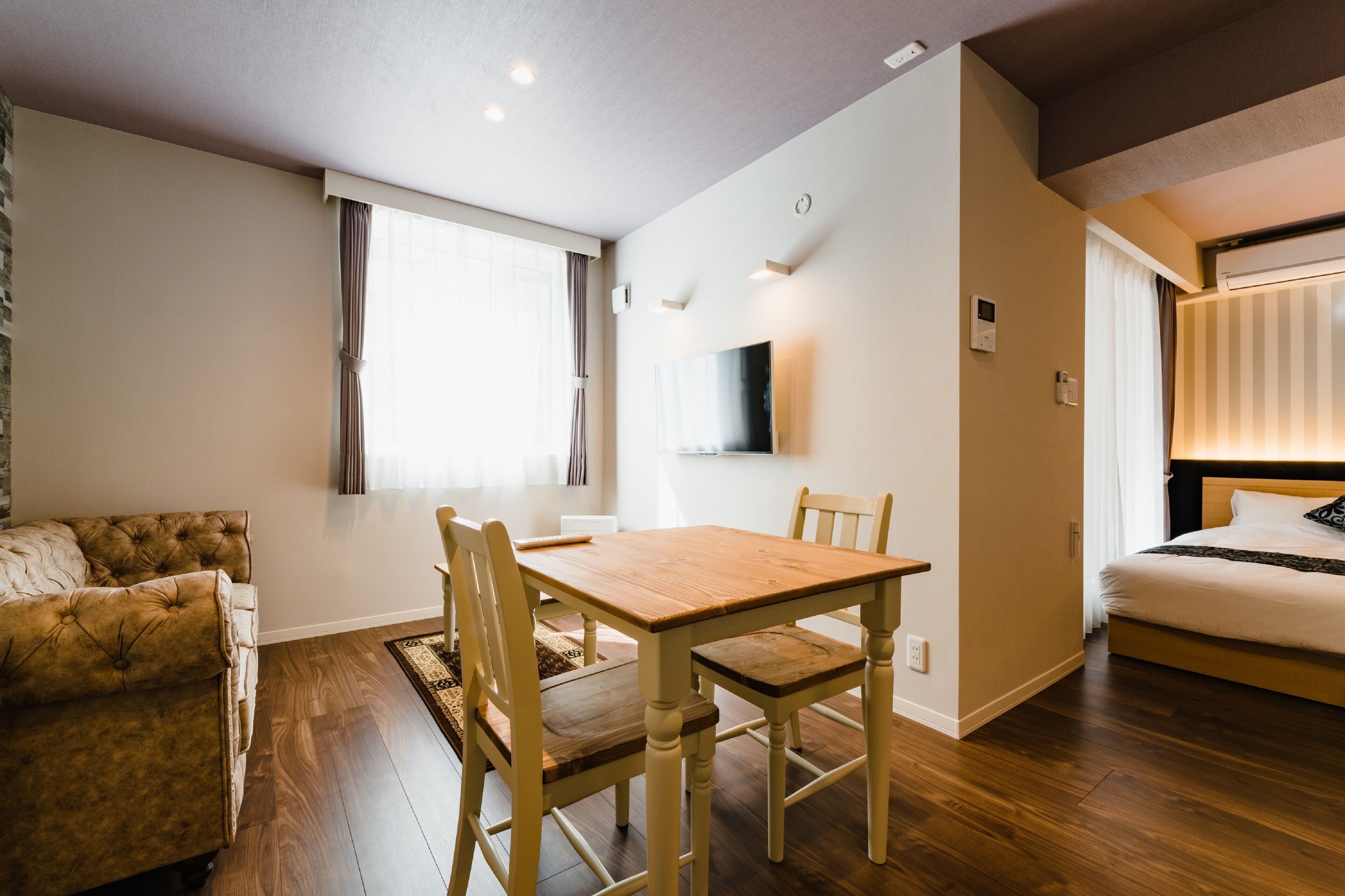 A room with a wooden dining table, two chairs, a sofa, a bed, and a TV mounted on the wall.
