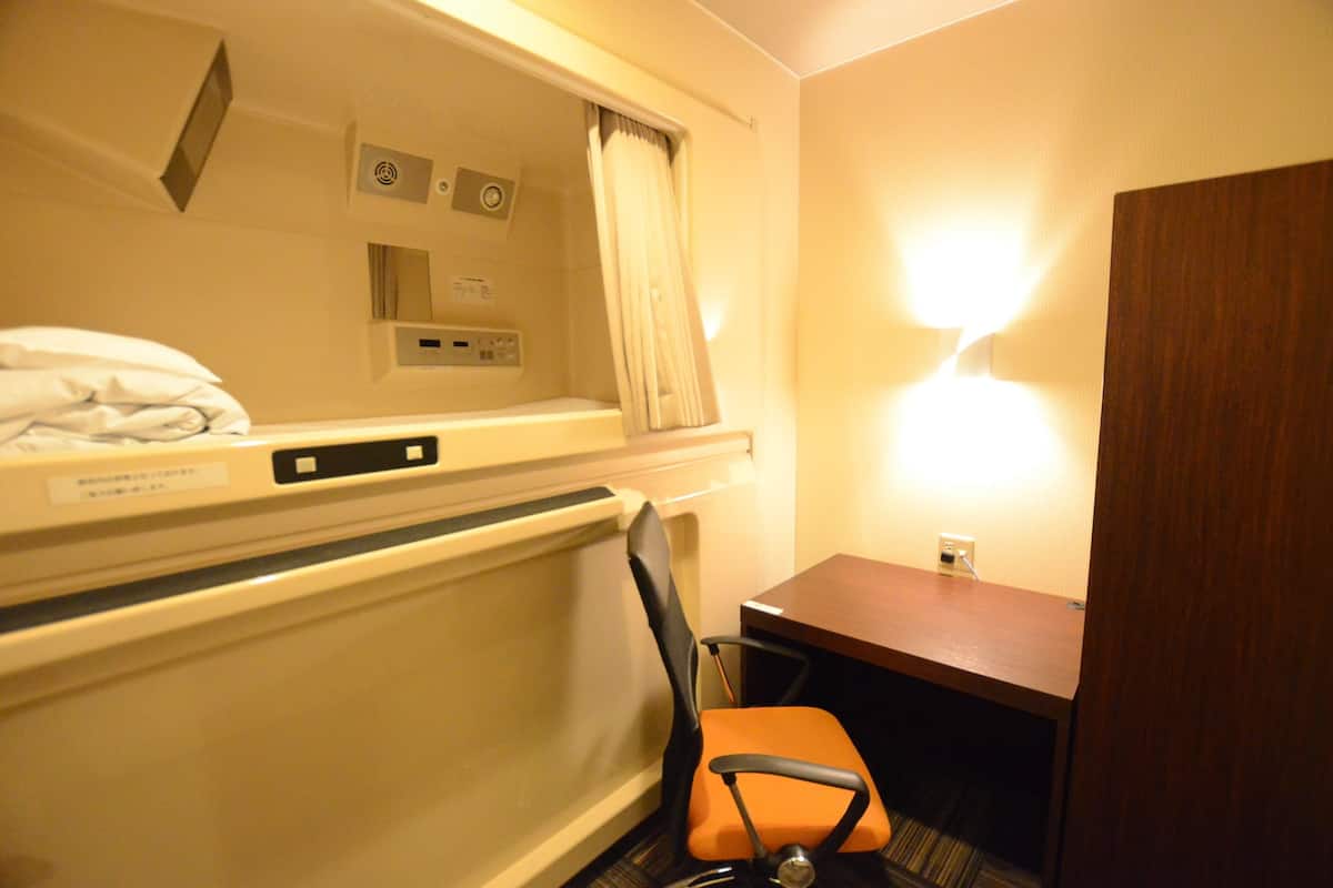 Ein Hotelzimmer mit einem Bett, einem Schreibtisch, einem orangefarbenen Sessel, einer Holztür und einer Wandleuchte.