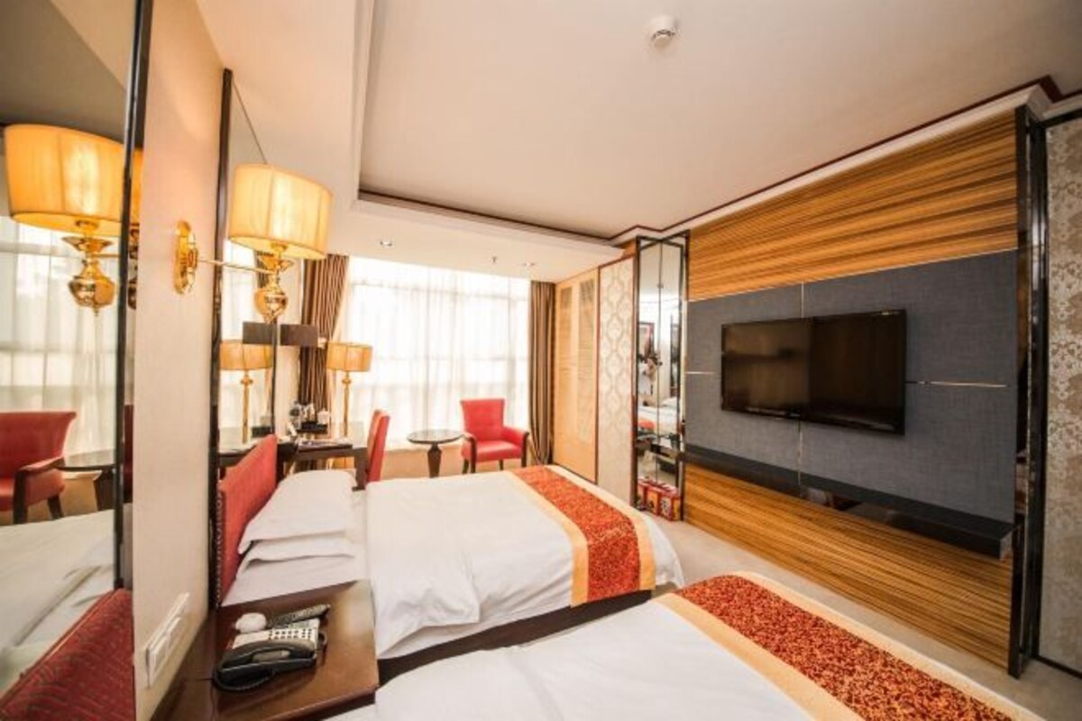 A hotel room with two beds, a TV, a desk with a chair, and a small table with a lamp.