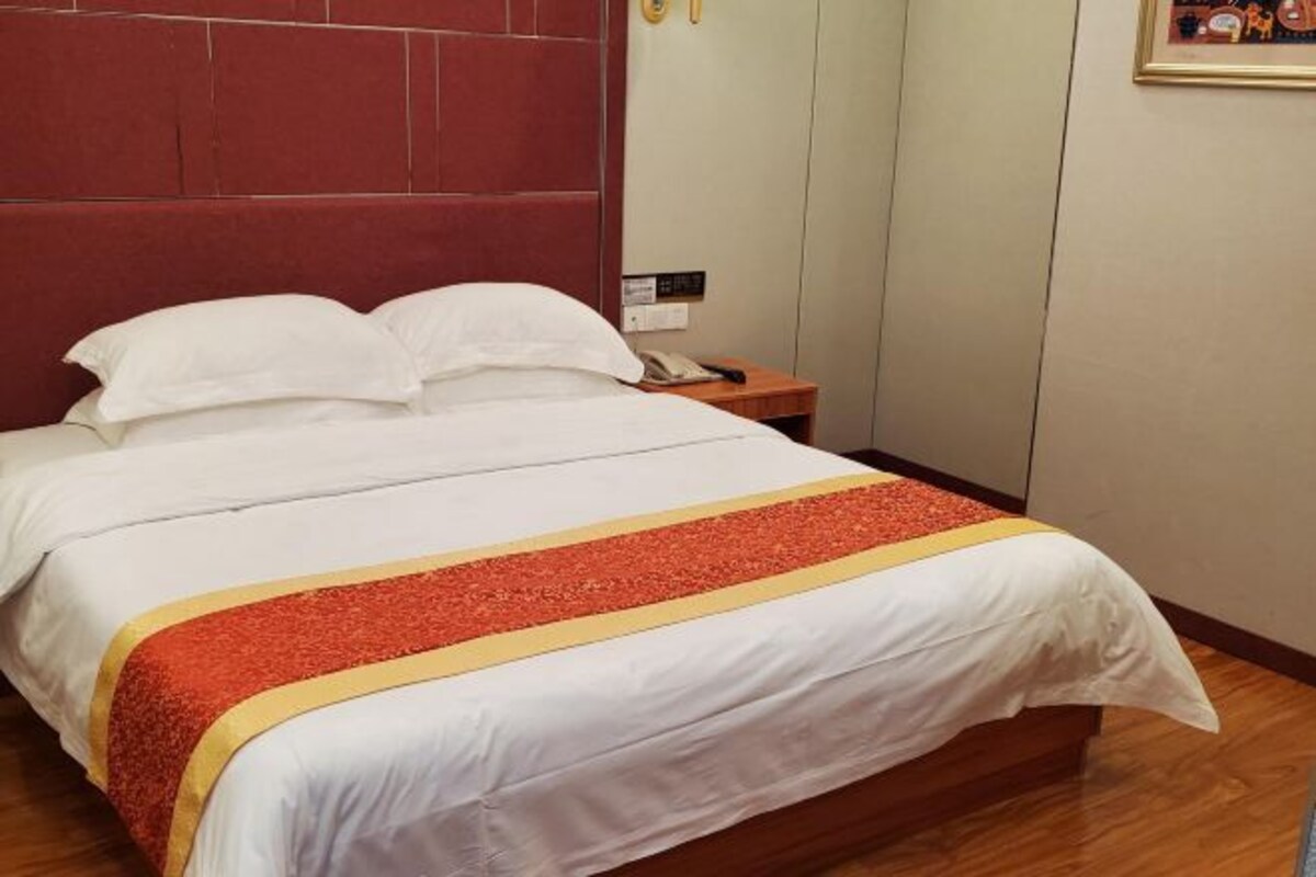 A hotel room with a bed, a wooden headboard, a nightstand with a phone, and a wall-mounted light switch.
