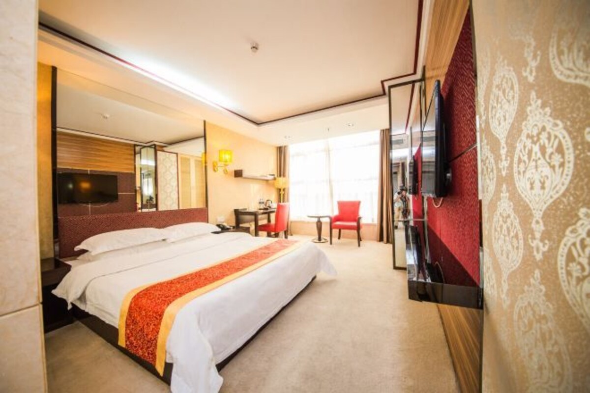 A hotel room with a bed, a TV mounted on the wall, a desk with a chair, and a large window.