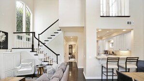Amplia sala de estar con escalera, cocina con muebles blancos y comedor con sillas.