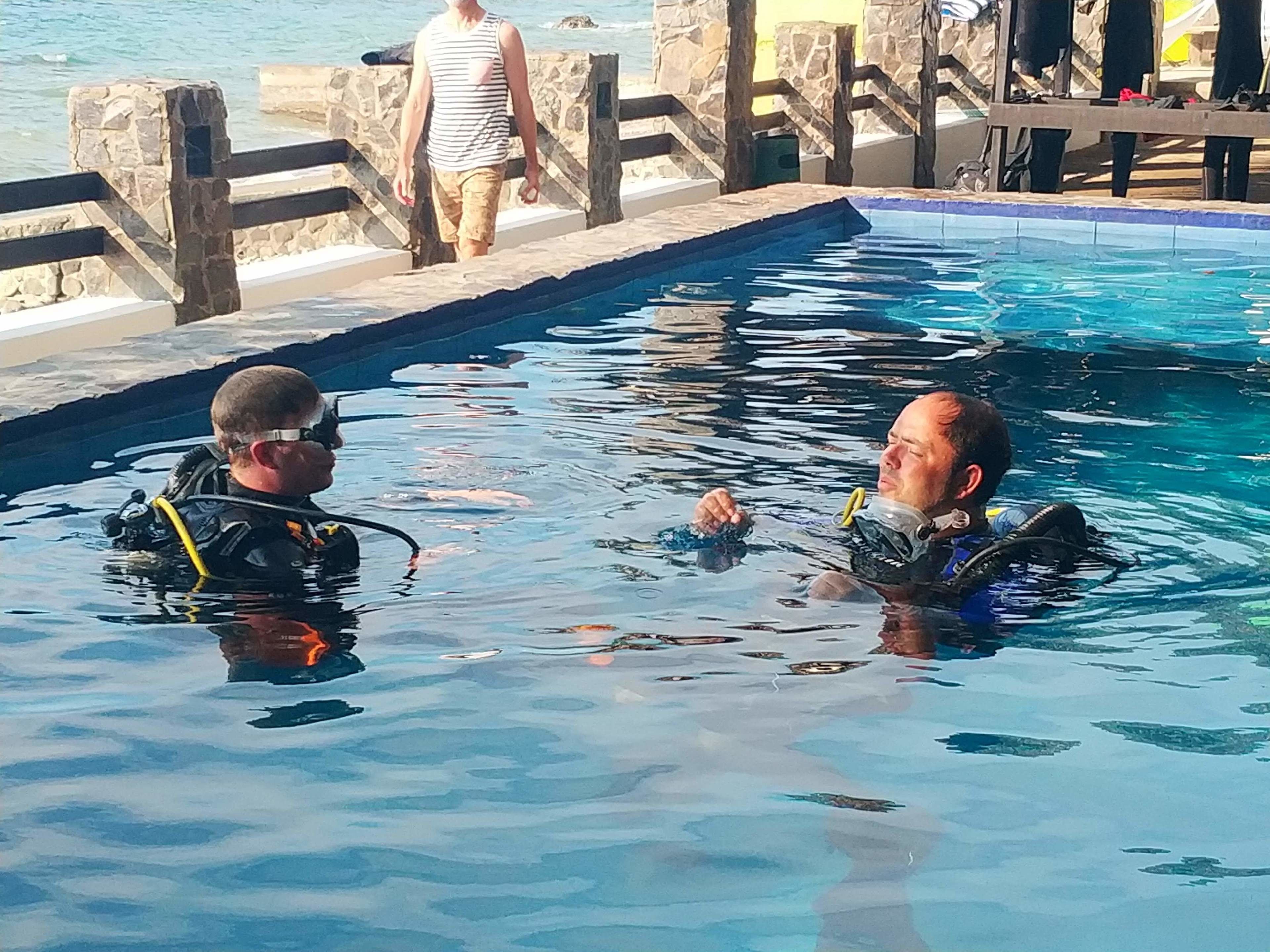 Dos personas en una piscina, con una pared de piedra y una terraza de madera al fondo.