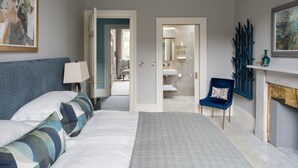 Ein Schlafzimmer mit Bett, Sessel, Lampe, Kamin und einem durchgehenden Türspalte sichtbaren Badezimmer.