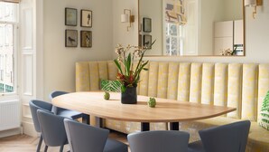 Ein Esszimmer mit einem runden Tisch, sechs Stühlen, einer Bank mit gelb gemusterten Kissen und einem großen Spiegel an der Wand.