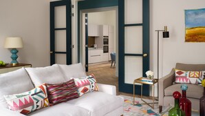 Ein Wohnzimmer mit einem weißen Sofa, bunten Kissen, einem Couchtisch mit dekorativen Flaschen und Blick in die Küche durch offene Türen.