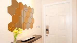 A hallway with a white door, a wall-mounted hexagonal mirror, a dark shelf with a vase of yellow flowers, and a small rectangular object on the shelf. - Fv62884 - Regal Oaks - 4 Bed 3.5 Baths Townhome (Kissimmee)