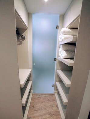 Un couloir étroit, avec une porte au fond, bordé d’étagères où sont pliés des draps et autres articles de literie.