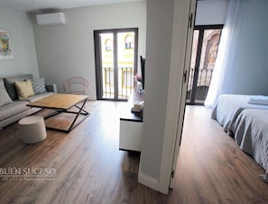 Un salon moderne avec un canapé gris, une table basse en bois et un fauteuil rouge. Un balcon offre une vue sur un immeuble et une statue.