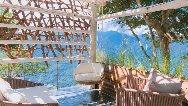 Une terrasse meublée de mobilier en osier, avec une chaise blanche, et une vue sur les montagnes et les arbres.