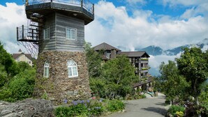 Une tour de pierre dotée d’un balcon, entourée d’une végétation luxuriante et avec une chaîne de montagnes en toile de fond.