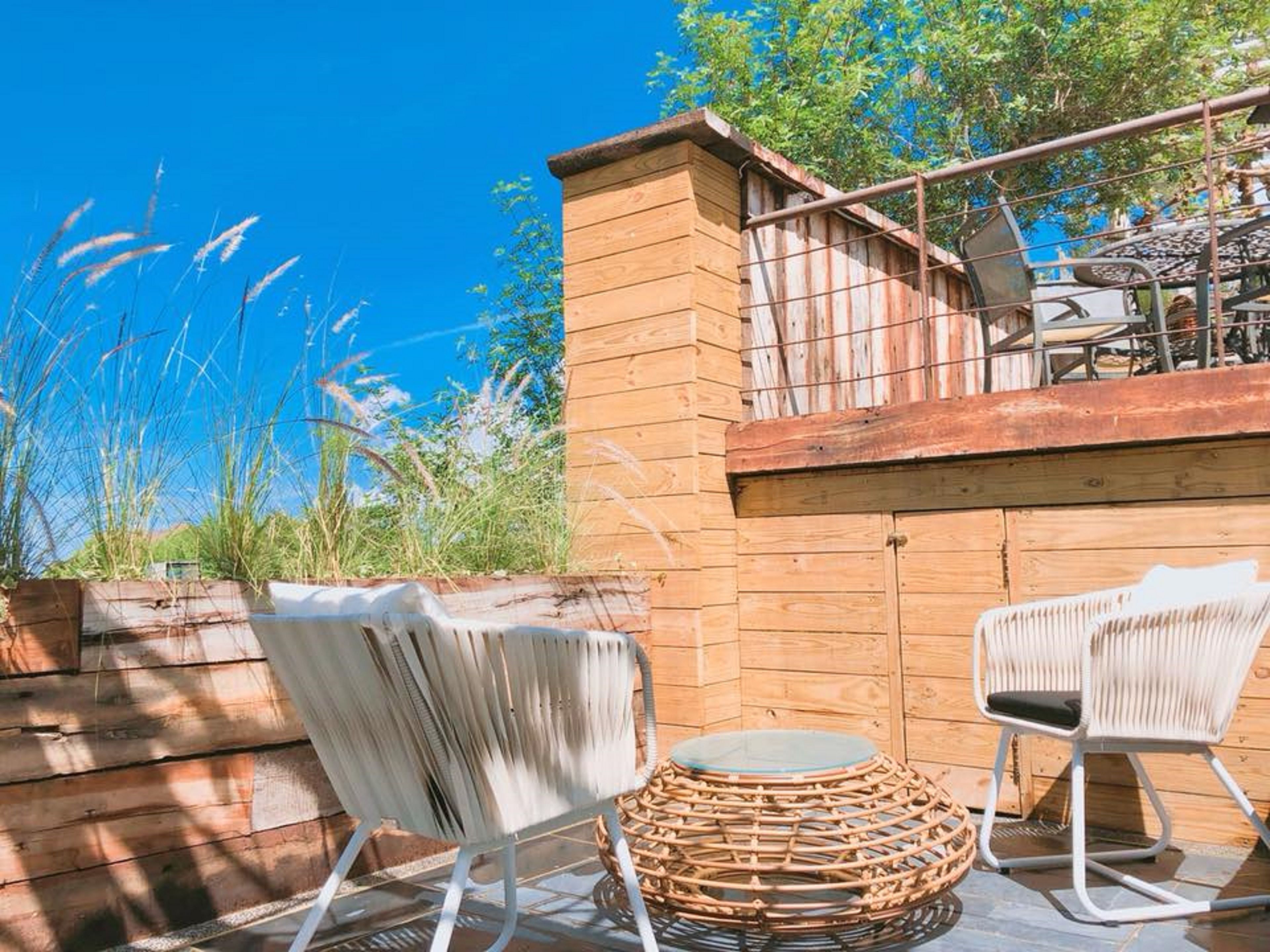 A wooden deck with two wicker chairs and a round table, overlooking a garden with tall grasses and trees.