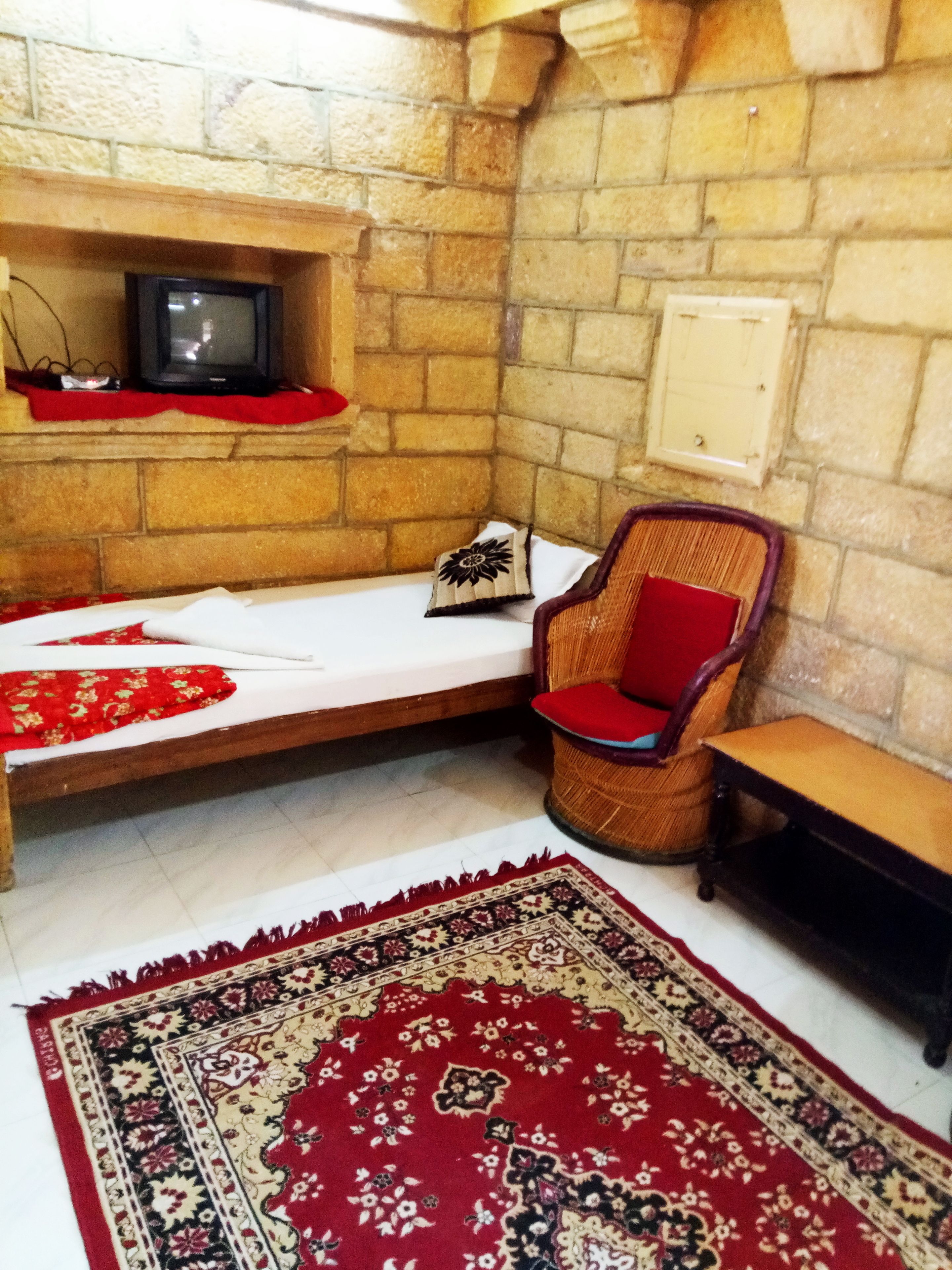 Una habitación con pared de piedra, cama de madera, silla tejida, una mesita y un tapete con diseño rojo y negro.
