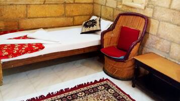 Una habitación con pared de piedra, cama de madera, silla tejida, una mesita y un tapete con diseño rojo y negro.