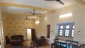 A spacious dining area with wooden tables and chairs, ceiling fans, and a large window with decorative shutters. - Hotel Paradise (Jaisalmer)