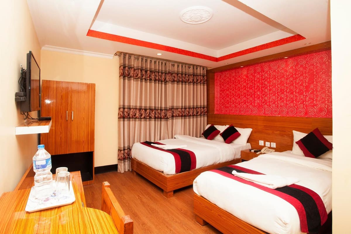 Habitación de hotel con dos camas, un armario de madera, un televisor, una mesa con un recipiente de agua y una pared con un estampado rojo.