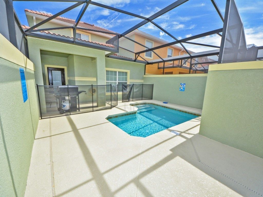 An outdoor swimming pool with a glass enclosure, surrounded by a light-colored patio area and a metal roof structure.