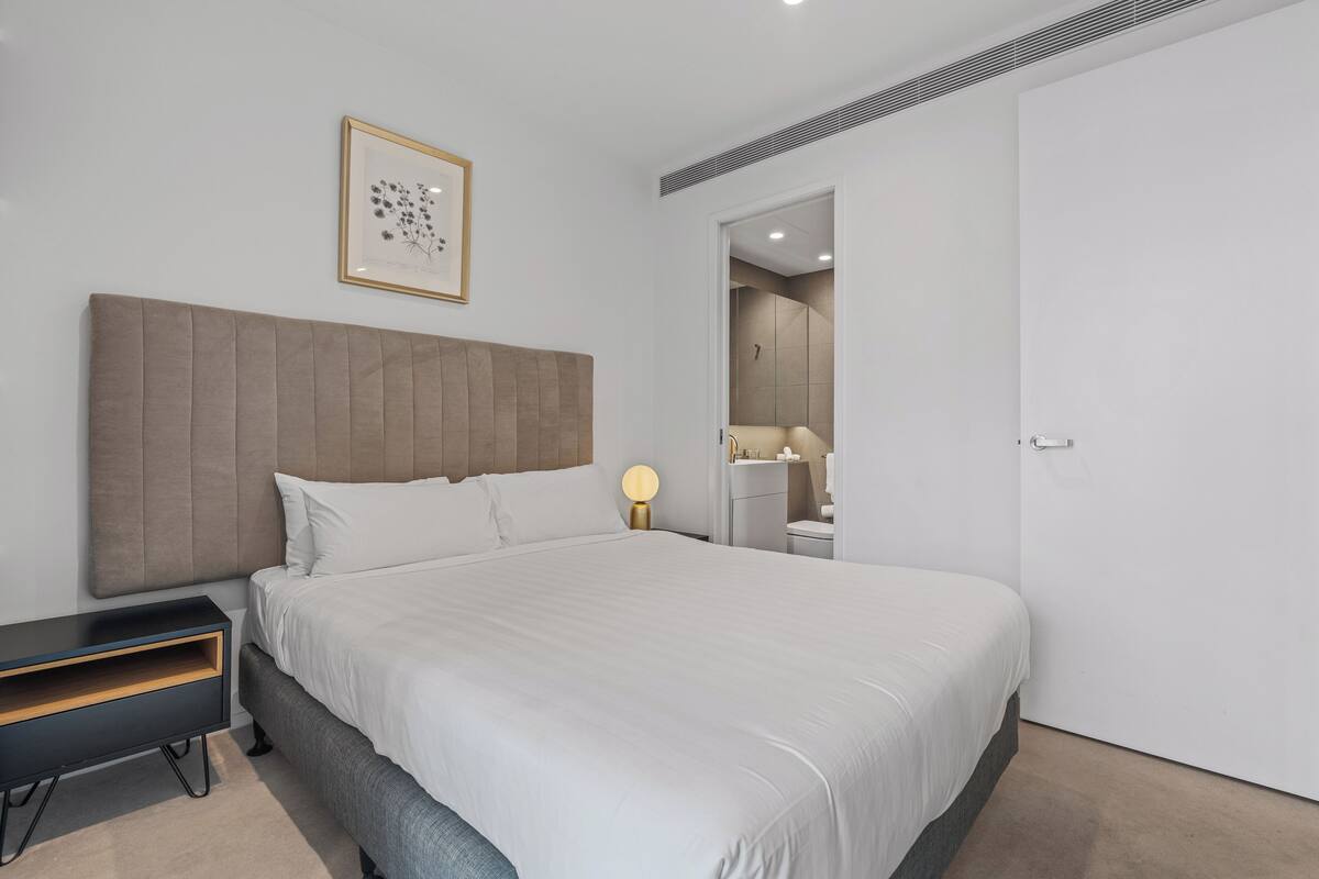 A modern bedroom with a large bed, a bedside table, a framed picture on the wall, and a door leading to a bathroom.