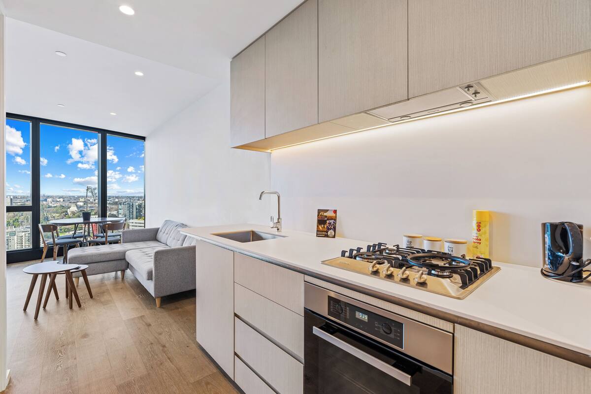 A modern kitchen with a built-in stove, oven, and sink. There is a dining area with a gray sofa and a small table. The kitchen has large windows offering a city view.