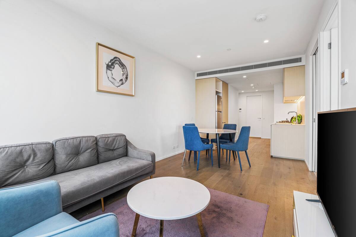 A modern living room with a grey sofa, a round white table, and a blue chair. There is a kitchen area with a white countertop and blue chairs.