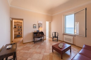 Een woonkamer met een open haard, een boekenkast, een eettafel en uitzicht op de zee door een raam.