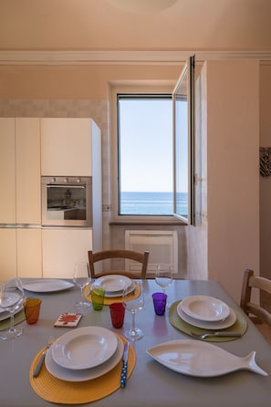 Een eetruimte met een tafel gedekt voor vier personen, uitzicht op zee door een raam, en een keuken met een inbouwoven.