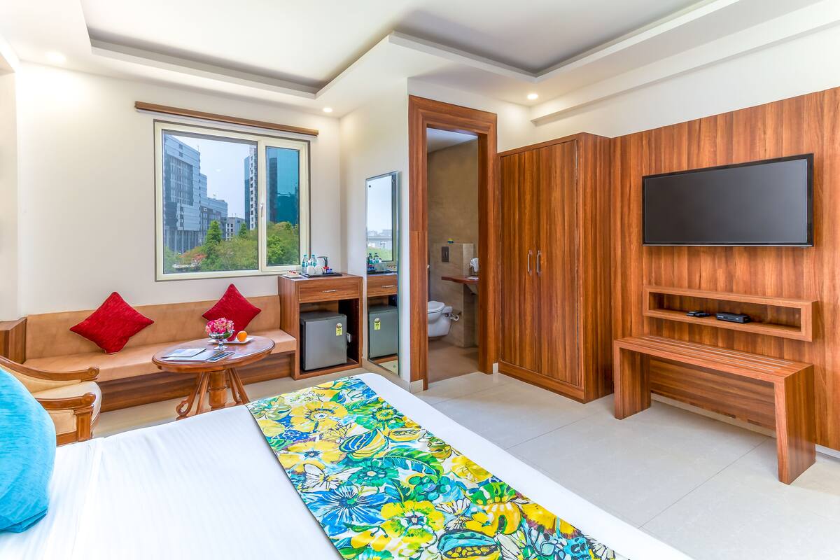 A modern hotel room with a large bed, a sofa, a small table, a TV mounted on a wooden wall, and a view of the city through a window.