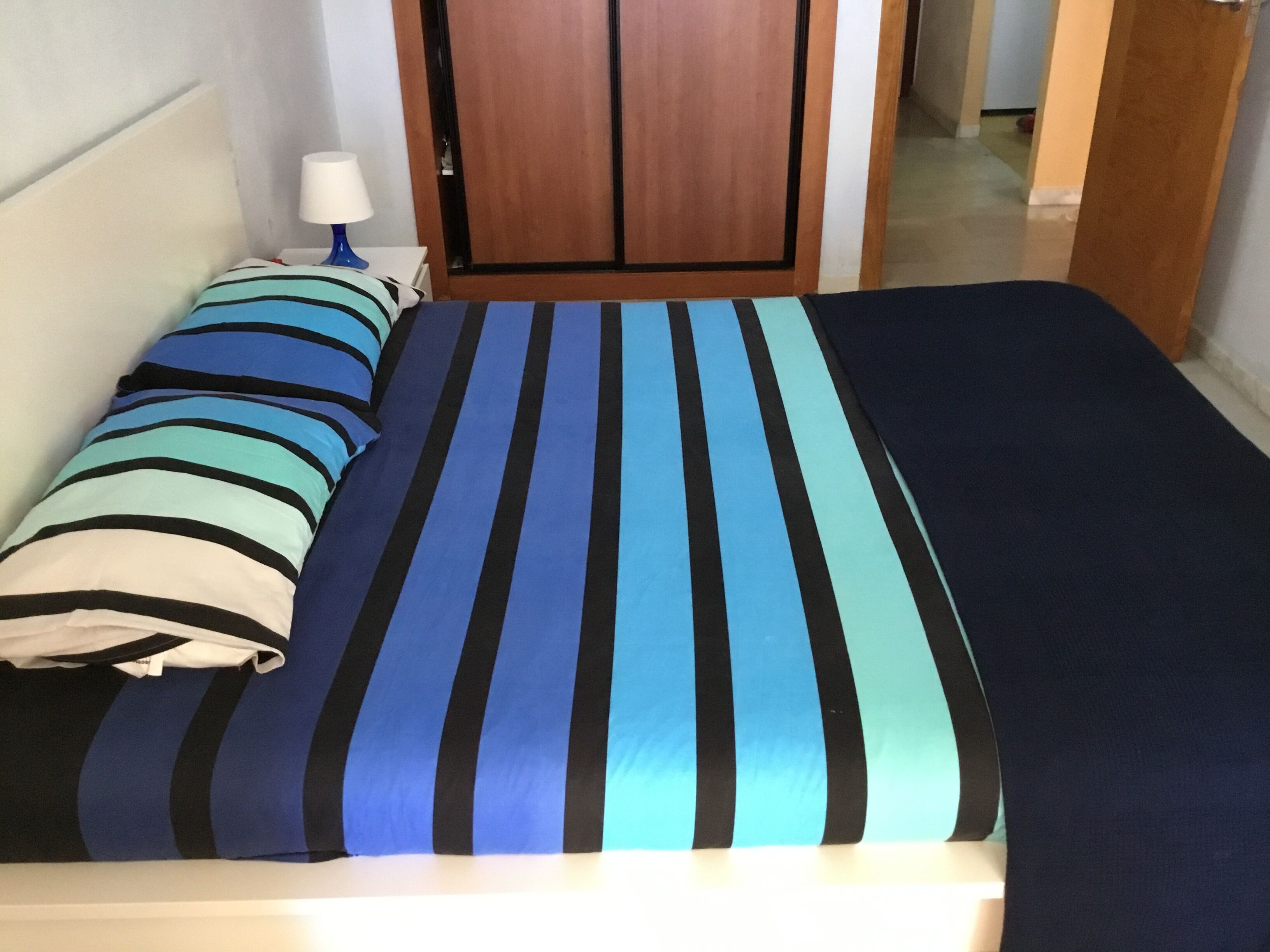 A bed with striped bedding in a room with a wooden door and a nightstand with a lamp.