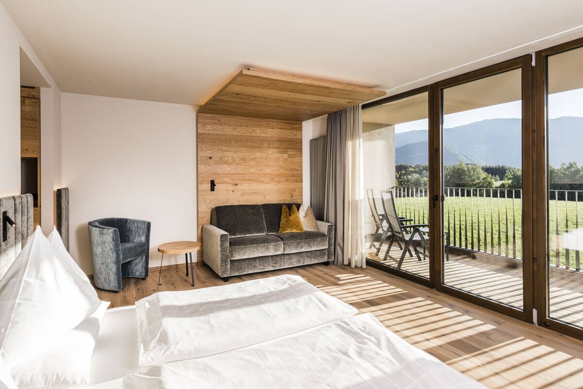 A modern bedroom with a large bed, a sofa, a small table, and a balcony with a view of the countryside.