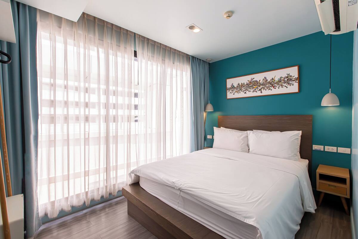 Una habitación de hotel moderna con una cama grande, cabecera de madera, pared de acento color aguamarina y un ventanal con cortinas translúcidas.