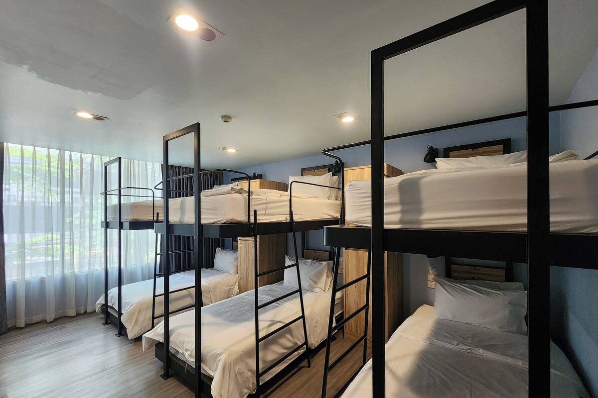 Habitación de hotel con literas, piso de madera y un ventanal con cortinas traslúcidas.