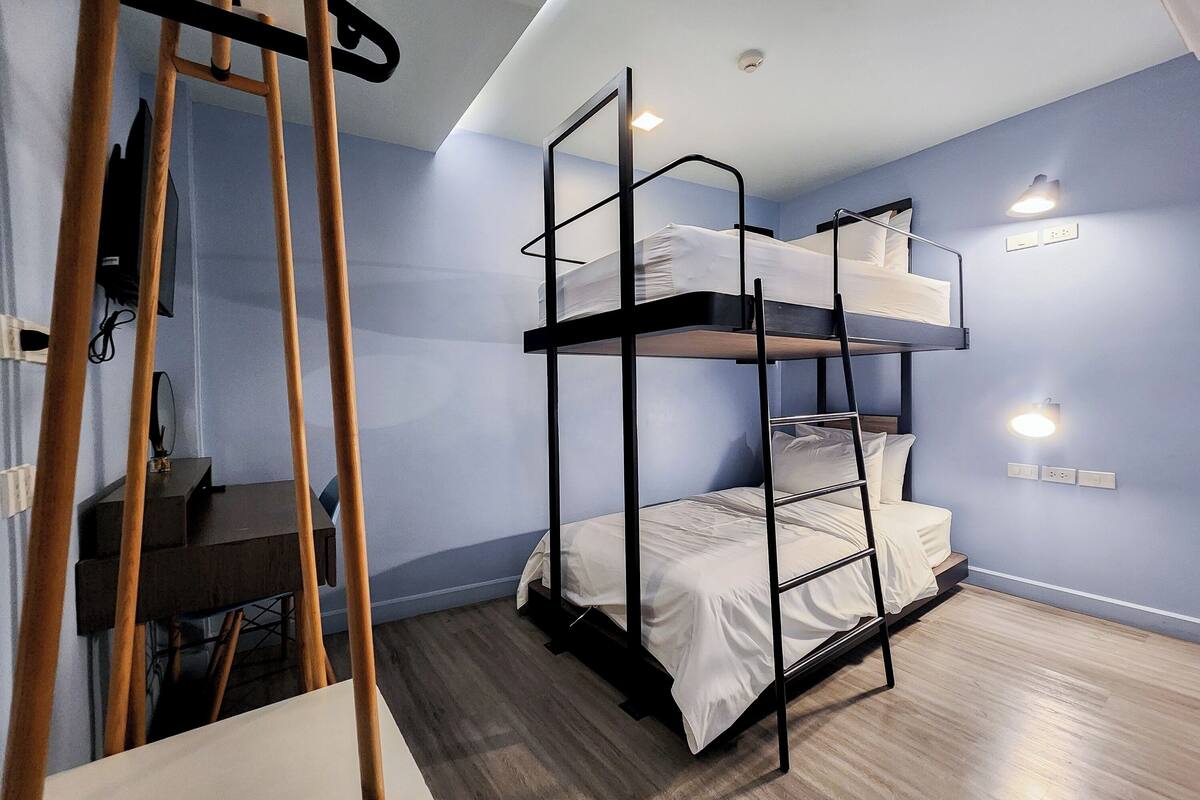 Dormitorio con literas, escritorio y escalera, suelo de madera y luces fijadas en la pared.