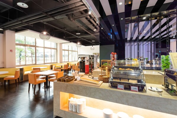 Un café moderno con zonas de estar, un mostrador de buffet con alimentos y amplias ventanas que dejan entrar luz natural.