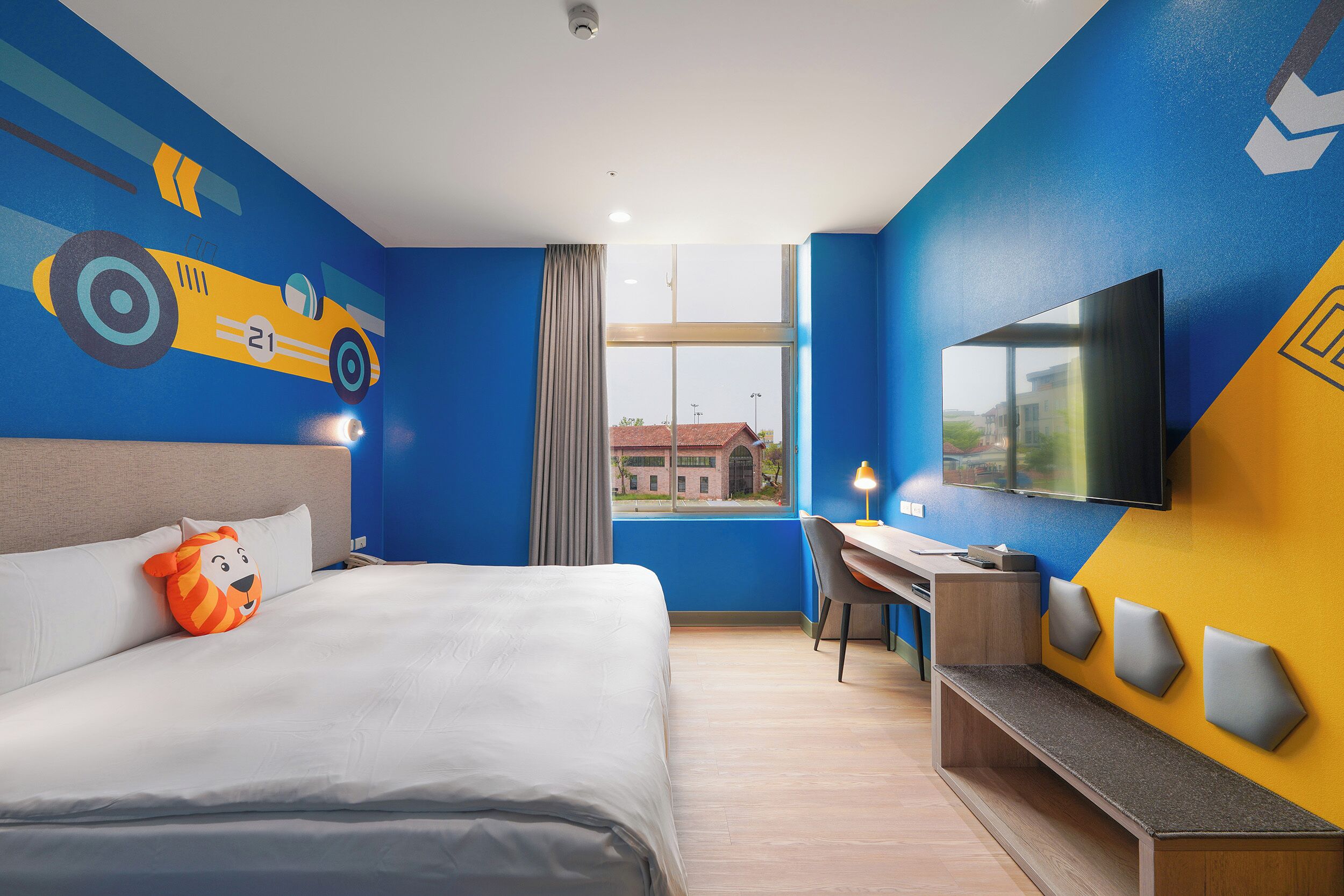 A hotel room with a bed, a desk, a TV, and a window with a view of buildings.