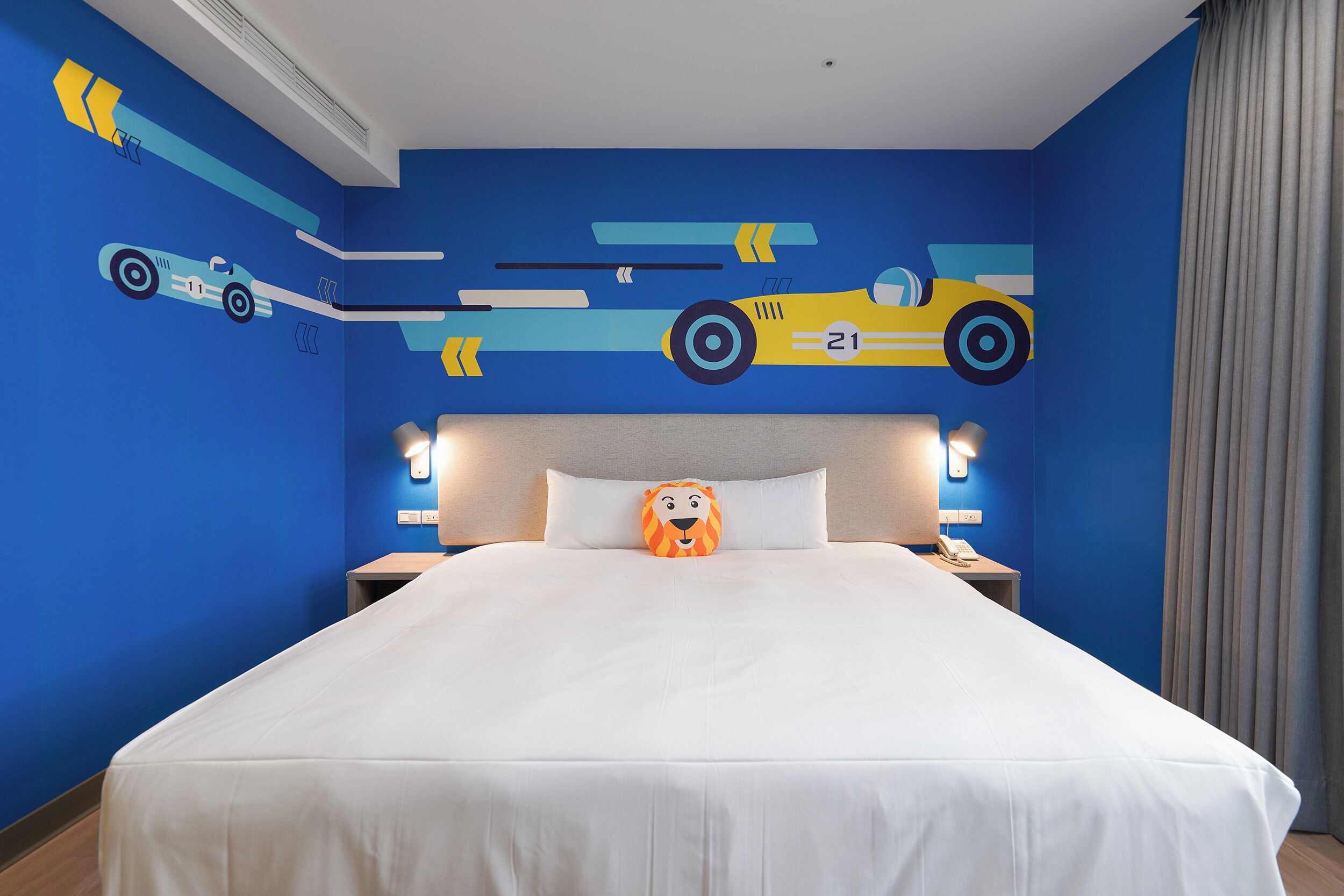 A hotel room with a large bed, a headboard with a light fixture, a nightstand with a lamp, and a wall with a race car mural.
