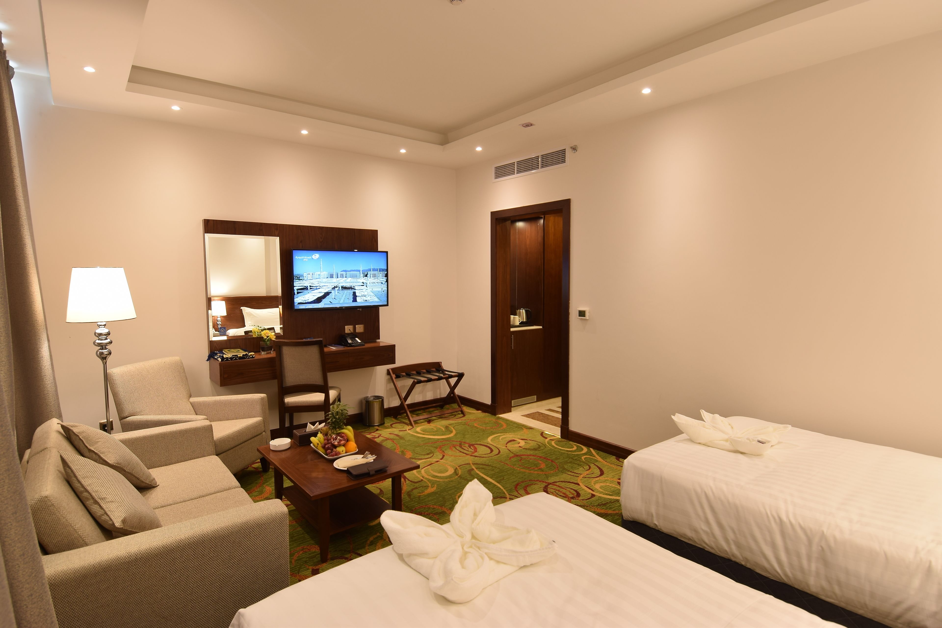 Hotelkamer met twee bedden, een bureau, een flatscreen-tv, een stoel, een lamp en een klein tafeltje met fruit en een fles.