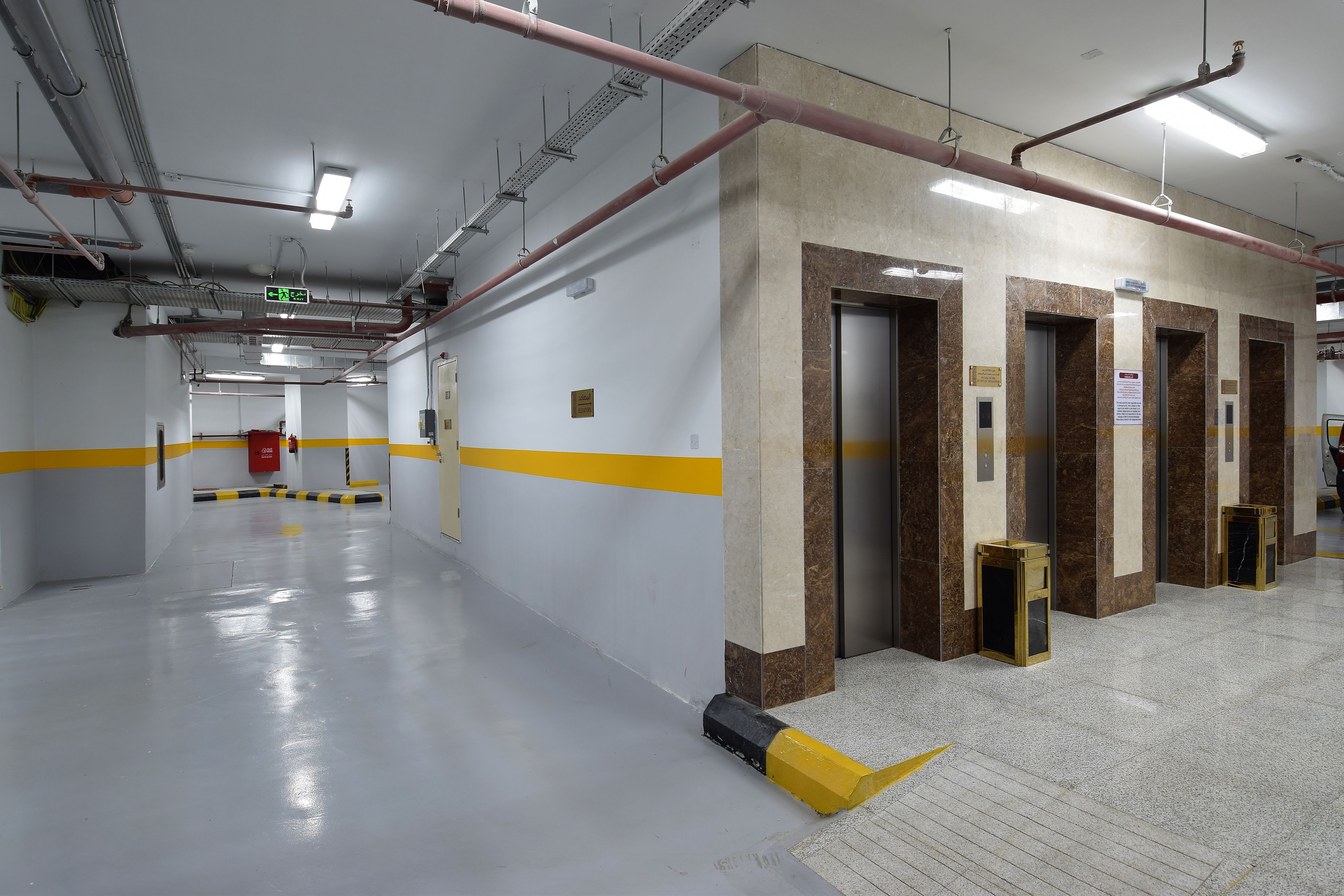 A clean, well-lit hallway with marble walls and yellow safety lines, leading to elevators and a fire extinguisher.