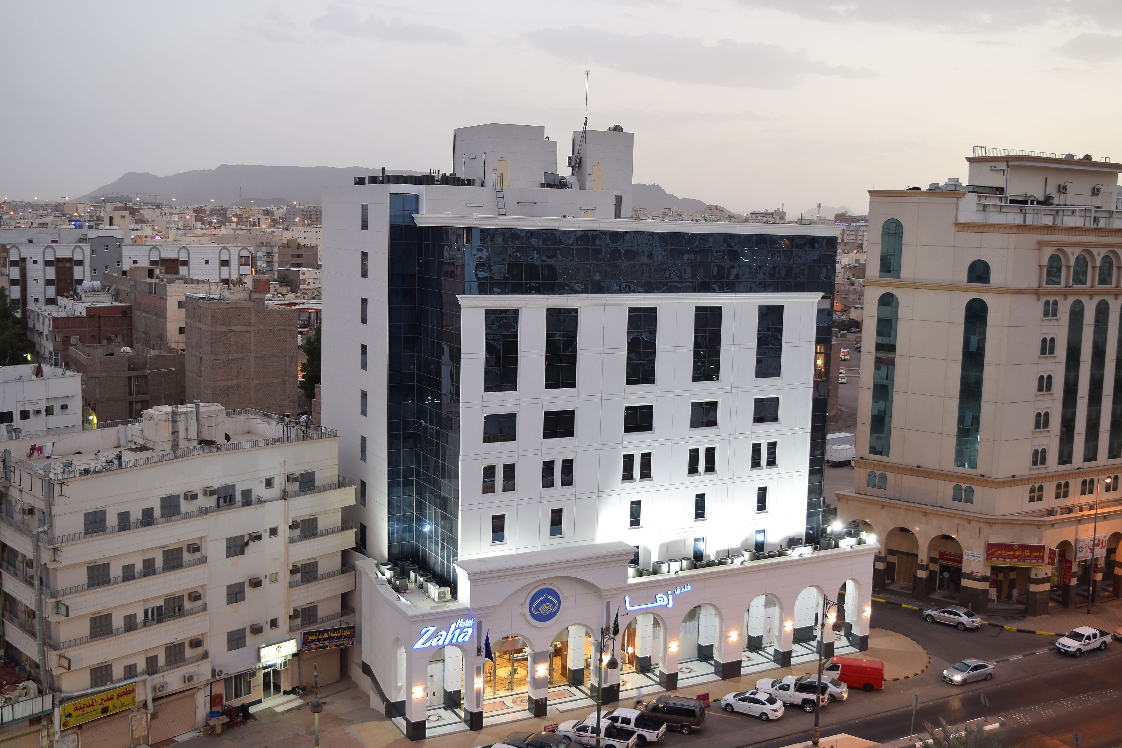 A hotel with a modern design, featuring a prominent entrance with a blue logo, multiple floors, and a parking area in front.