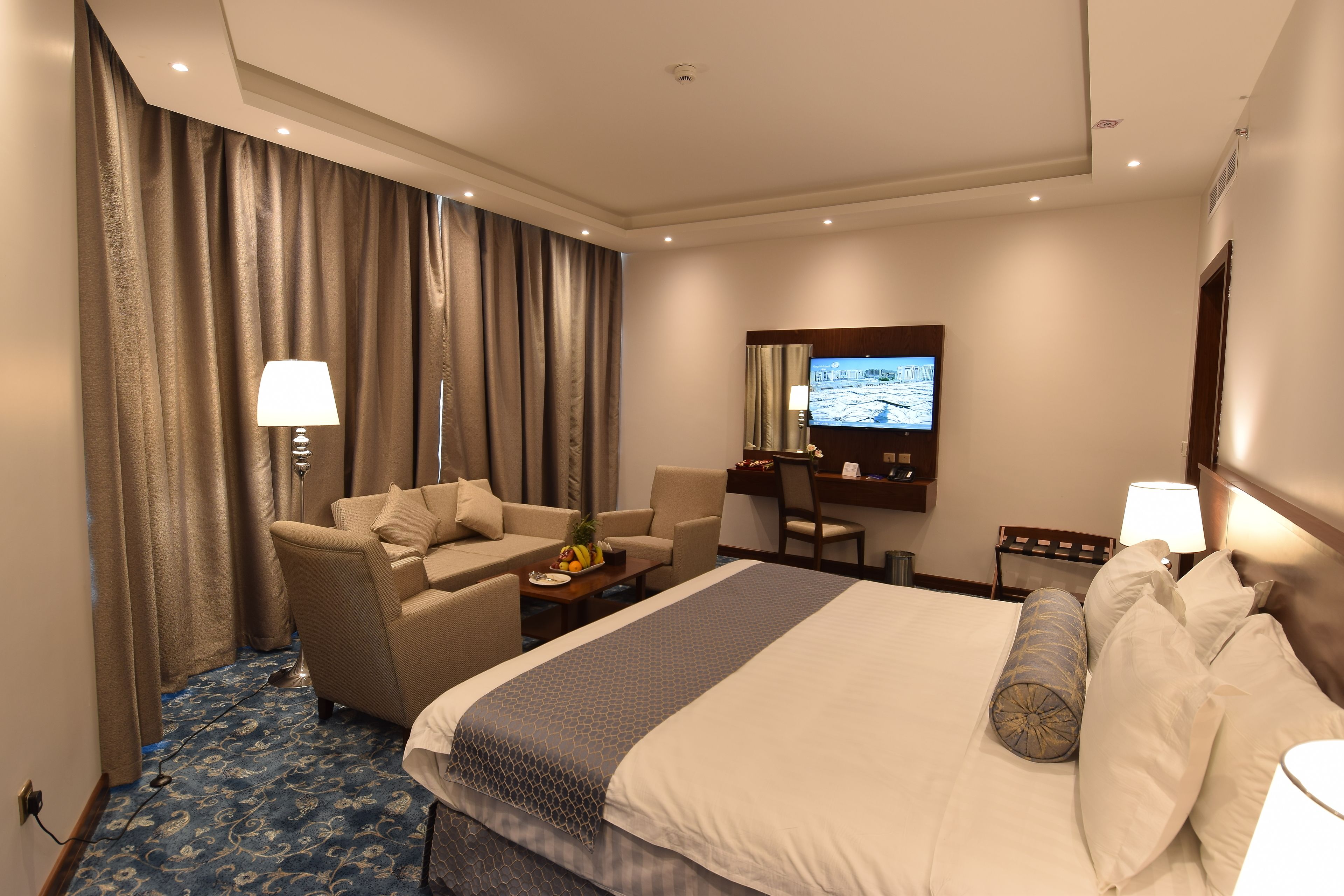 A hotel room with a large bed, a desk, a chair, a sofa, and a television.