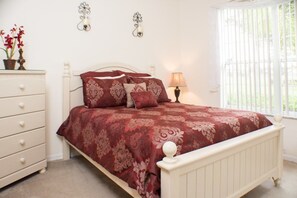 A bedroom with a white bed frame, a red and beige patterned bedspread, a white dresser with drawers, two wall sconces, and a window with blinds.