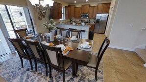 A dining area with a large table set for six, surrounded by wooden chairs. A kitchen with wooden cabinets and stainless steel appliances is visible in the background. - Aco240235 - Champions Gate Resort - 6 Bed 6 Baths Hotel (Davenport)