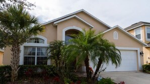 A residential house with a beige exterior, a white garage door, and a well-maintained garden with palm trees and red flowers. - Ip64290 - Seasons - 4 Bed 3 Baths Villa (Kissimmee)