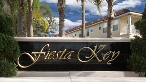 A sign with the name "Fiesta Key" in gold lettering on a black background, surrounded by palm trees and a building with a red-tiled roof.