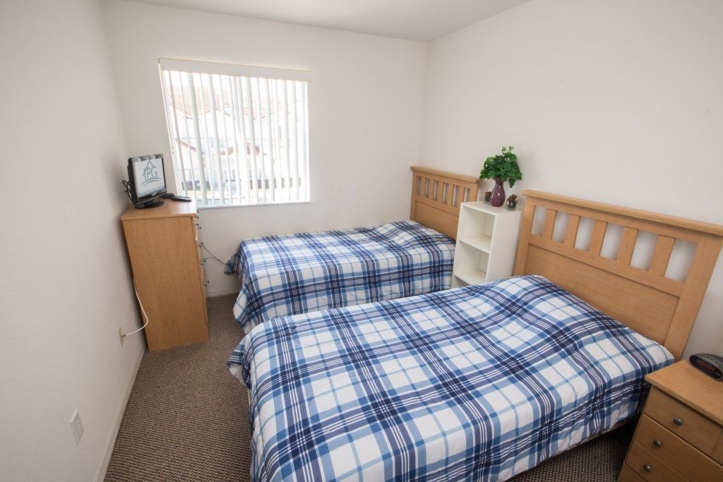 A bedroom with two beds, a wooden headboard, a small desk, a plant, and a window with blinds.