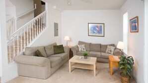 A living room with a beige sofa set, a wooden coffee table, and a small side table with a lamp. There is a staircase on the left and a framed picture on the wall. - Ip60350 - Fiesta Key - 3 Bed 2.5 Baths Townhome (Kissimmee)