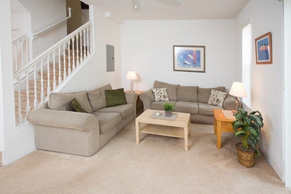 A living room with a beige sofa set, a wooden coffee table, and a small side table with a lamp. There is a staircase on the left and a framed picture on the wall.