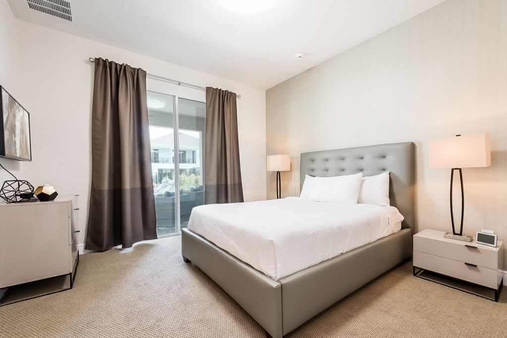 A modern hotel room with a large bed, two bedside lamps, a nightstand, a dresser, and a view through a glass door.