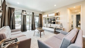 Una sala de estar moderna con sofá, sillón y una mesita de centro. Se aprecia un ventanal con cortinas, una estantería y una pequeña mesita auxiliar.